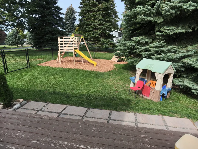 The fenced back yard with a wooden climbing frame, a slide and a playhouse on wood chips